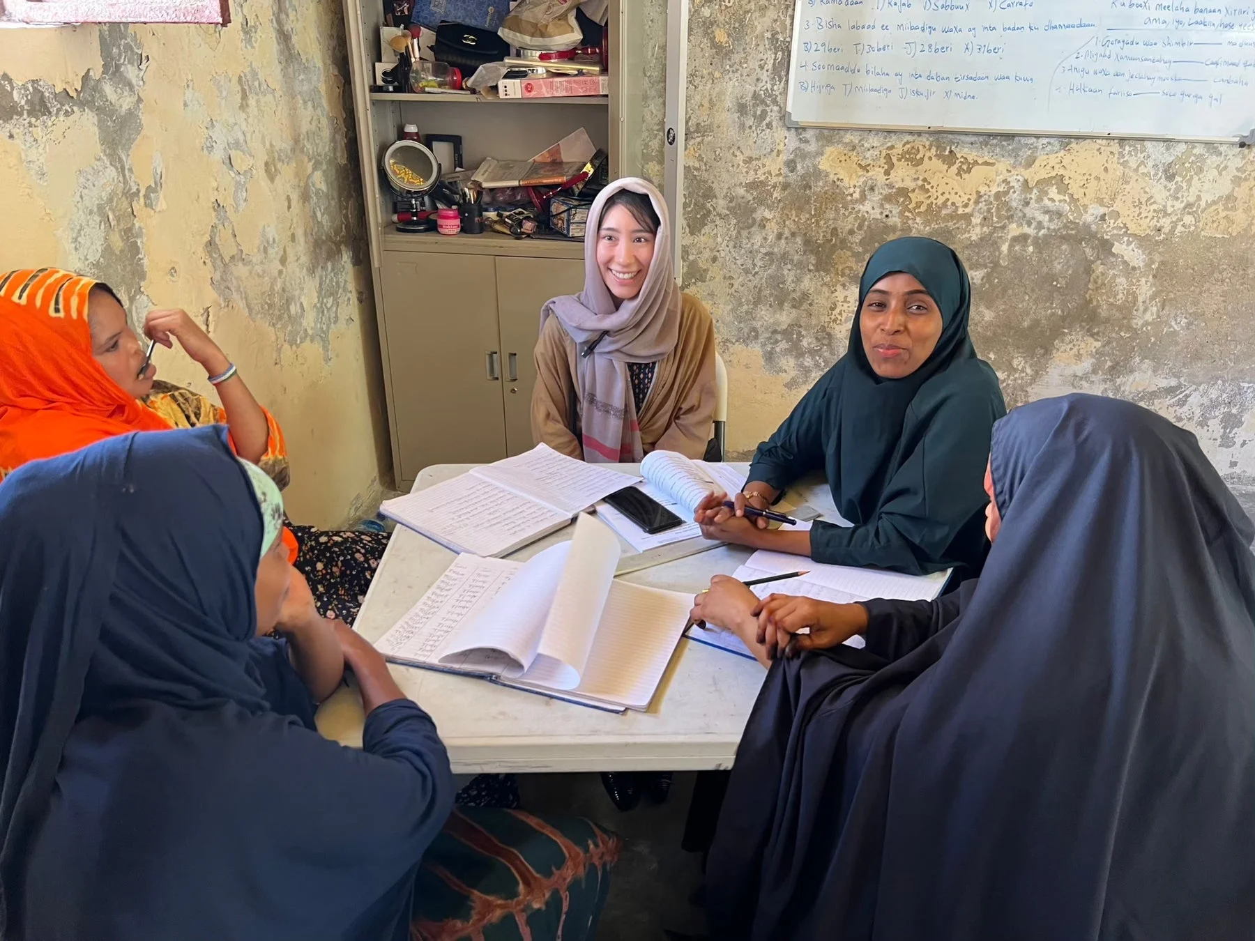 ヒジャブを着用した女性たちがテーブルを囲み、ノートやペンを使って学習している様子が写っています。笑顔を見せる女性もおり、簡素な教室で熱心にグループ学習が行われているようです。
