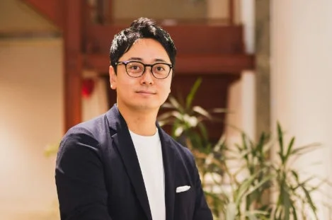 眼鏡をかけたアジア系の男性が、ネイビーのジャケットと白いTシャツを着用し、カメラに向かって微笑んでいるポートレート写真。背景はぼかされており、屋内の落ち着いた空間に見える。