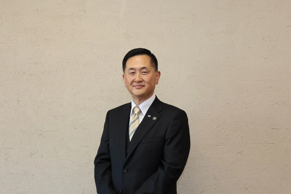 黒いスーツとストライプのネクタイを着用したアジア系の男性が、ベージュの壁を背景にカメラに向かって微笑んでいるポートレート写真。