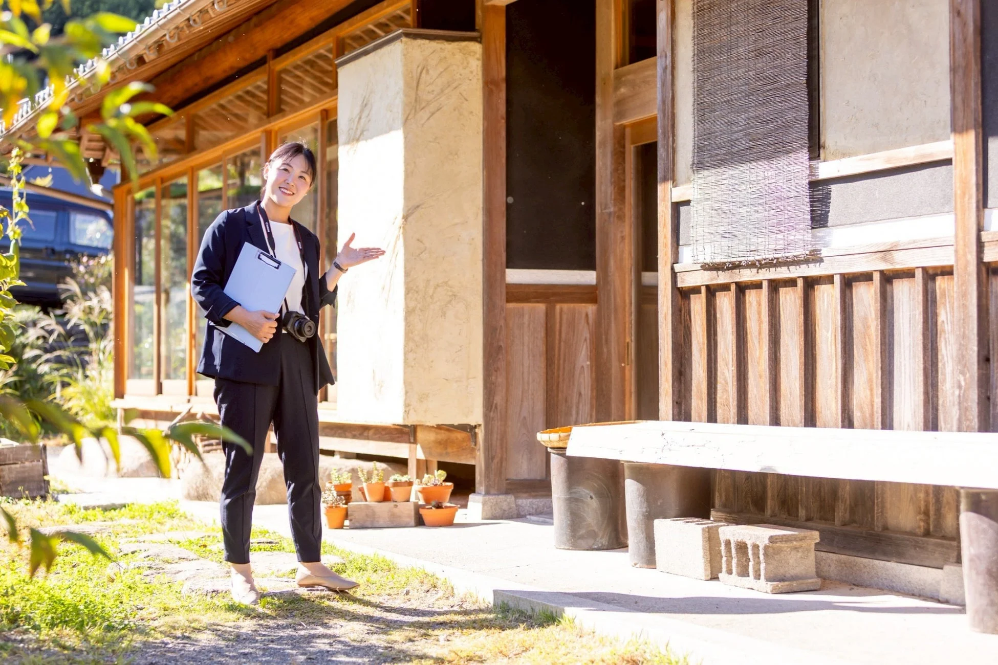 笑顔のアジア人女性が、和装の建物前でクリップボードとカメラを持ち、手で何かを指し示している