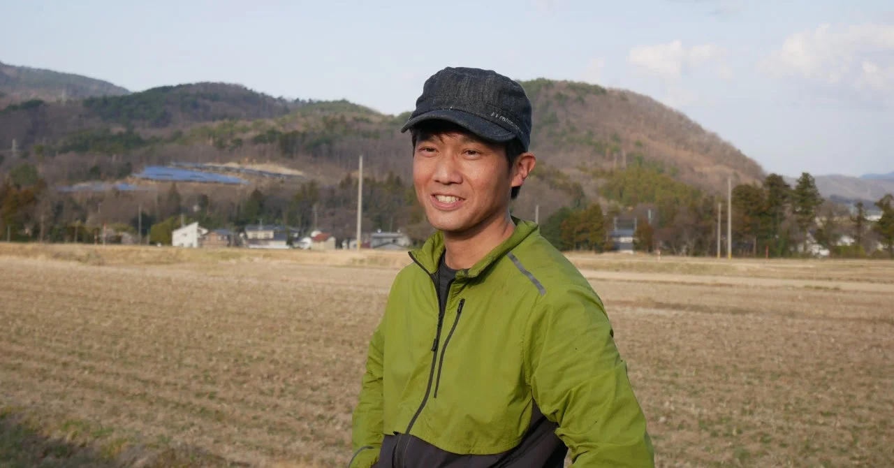 緑のジャケットとキャップを着用した男性が、乾燥した畑と山々を背景に笑顔で立っています