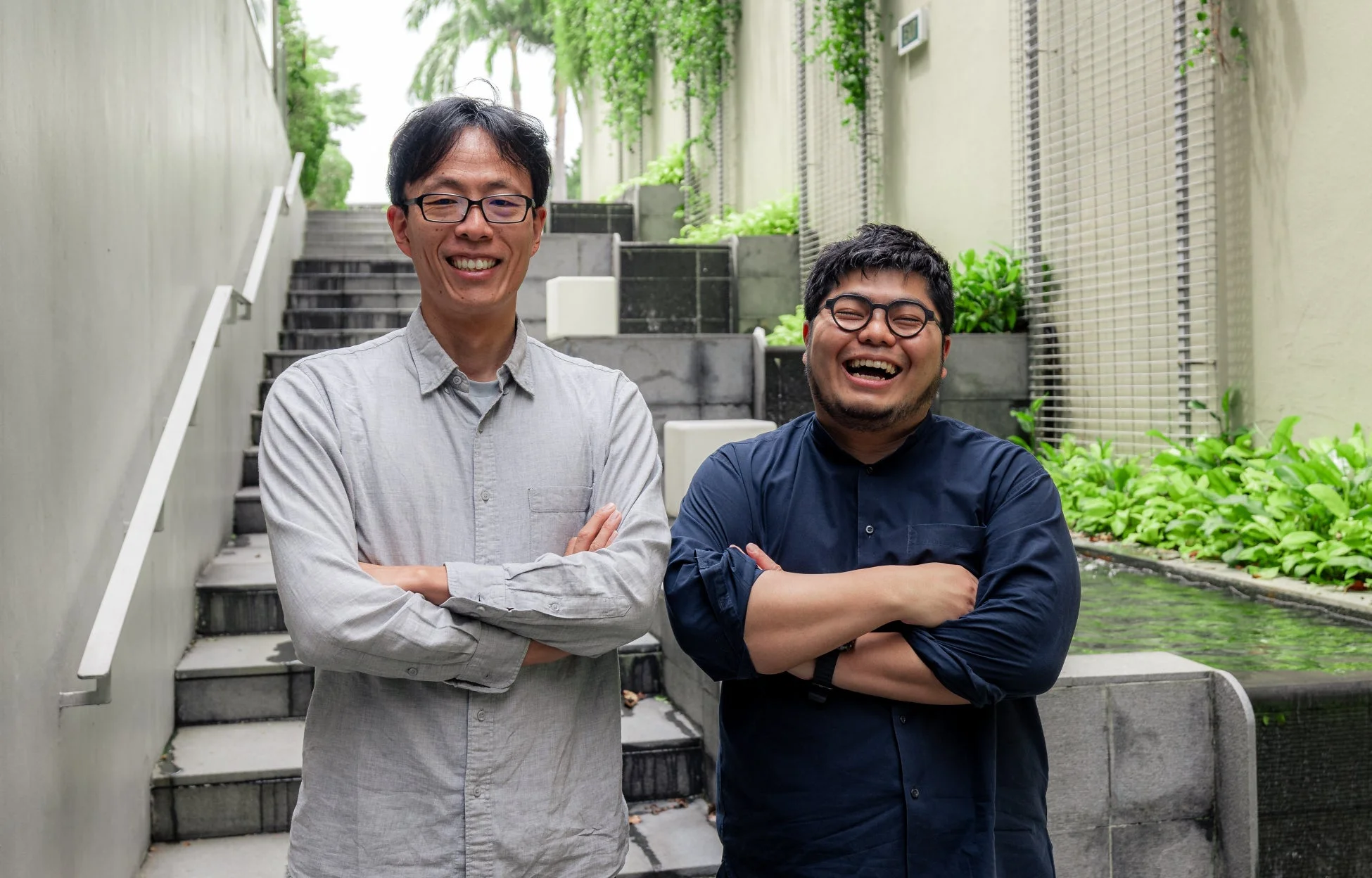 二人のアジア人男性が階段と緑豊かな背景の前で、腕を組みながら満面の笑みを浮かべているポートレート写真です。一人はグレーのシャツ、もう一人はネイビーのシャツを着ており、二人とも眼鏡をかけています。