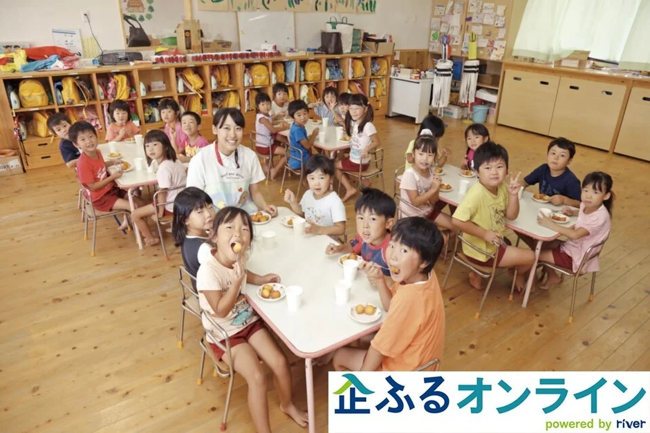 幼稚園か保育園の教室で、たくさんの園児たちが先生と一緒にテーブルを囲み、楽しそうにおやつを食べている様子。笑顔があふれ、明るい雰囲気です。