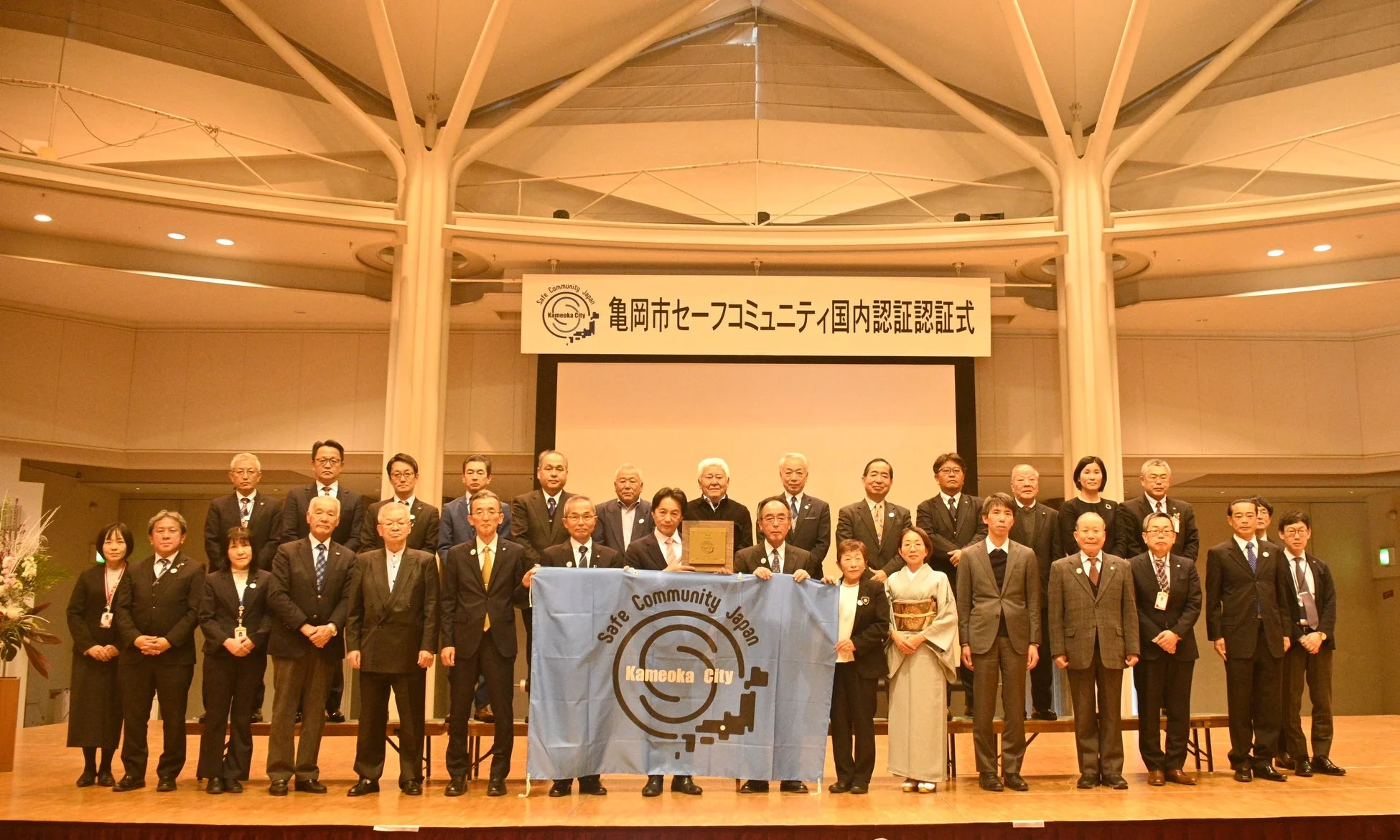 亀岡市セーフコミュニティの国内認証式典で、関係者がステージに集まり記念撮影をしている様子