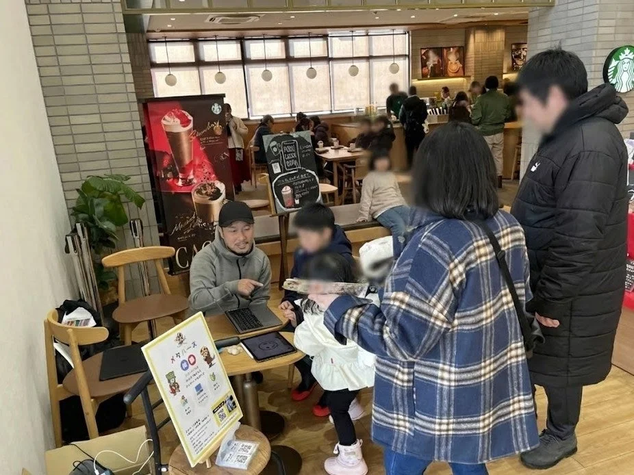 スターバックス店内で、多くの客が過ごしている賑やかな風景。手前では、男性が子どもたちにノートパソコンやタブレットを使って何かを説明しており、傍らには「メタバース」の体験会らしき案内板が立てられている