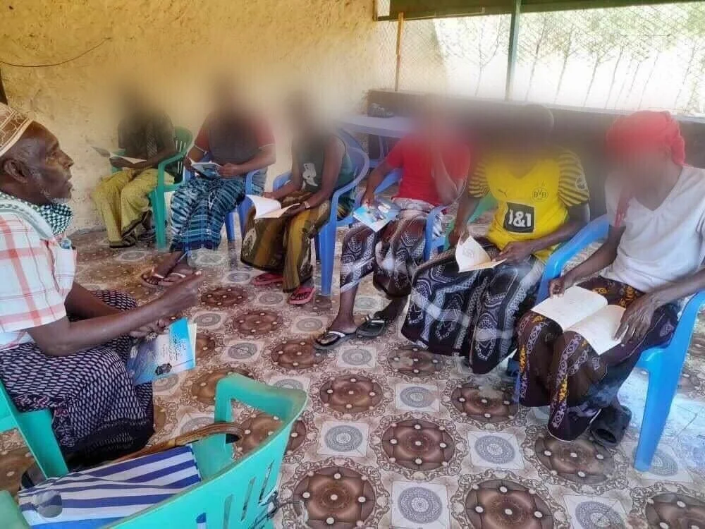 屋内で人々が椅子に座って集まり、年配の男性が話している様子です。参加者の中には本や書類を持っている人もおり、識字教室や地域での学習会、あるいは情報共有のための集まりであると推測されます。