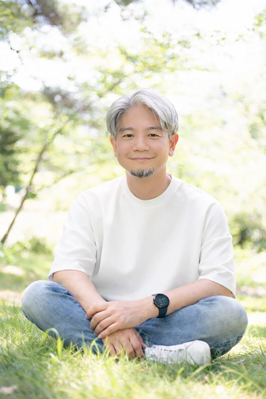 白髪とひげの男性が、明るい日差しの公園の芝生に胡坐をかいて座り、穏やかに微笑んでいます。