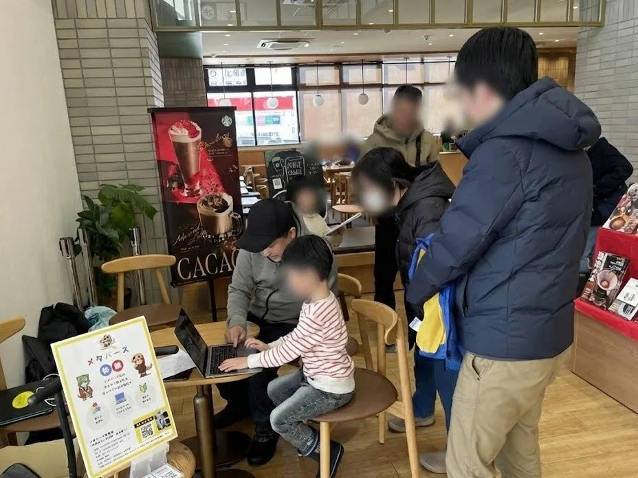 スターバックスのようなカフェで、父親と子供がノートパソコンを一緒に操作しています。隣には「メタバース体験」の案内板が立っており、デジタルコンテンツに触れる家族や人々で賑わいを見せています