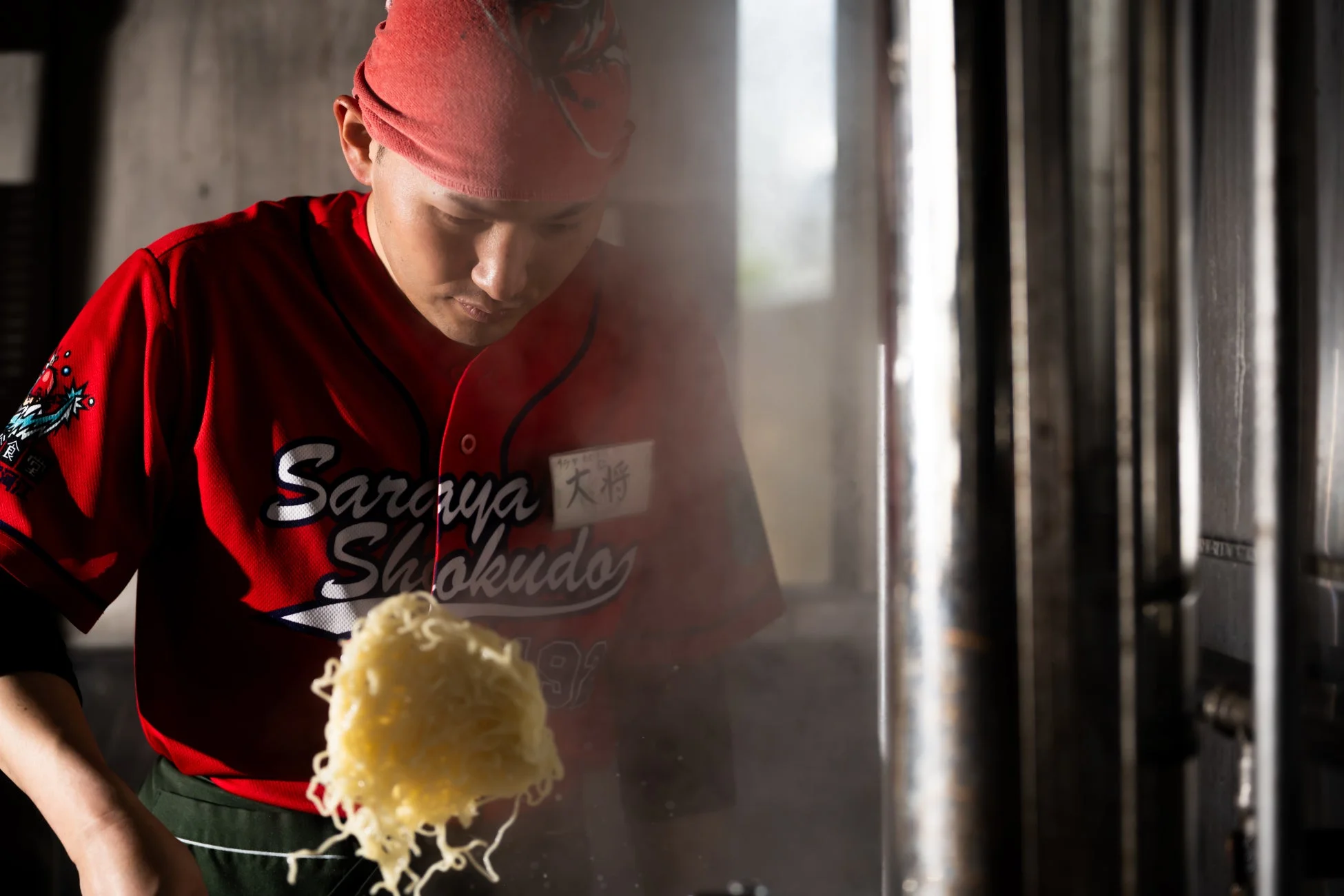 赤いユニフォームを着た料理人が、湯気の立つ厨房で麺を巧みに調理している様子。