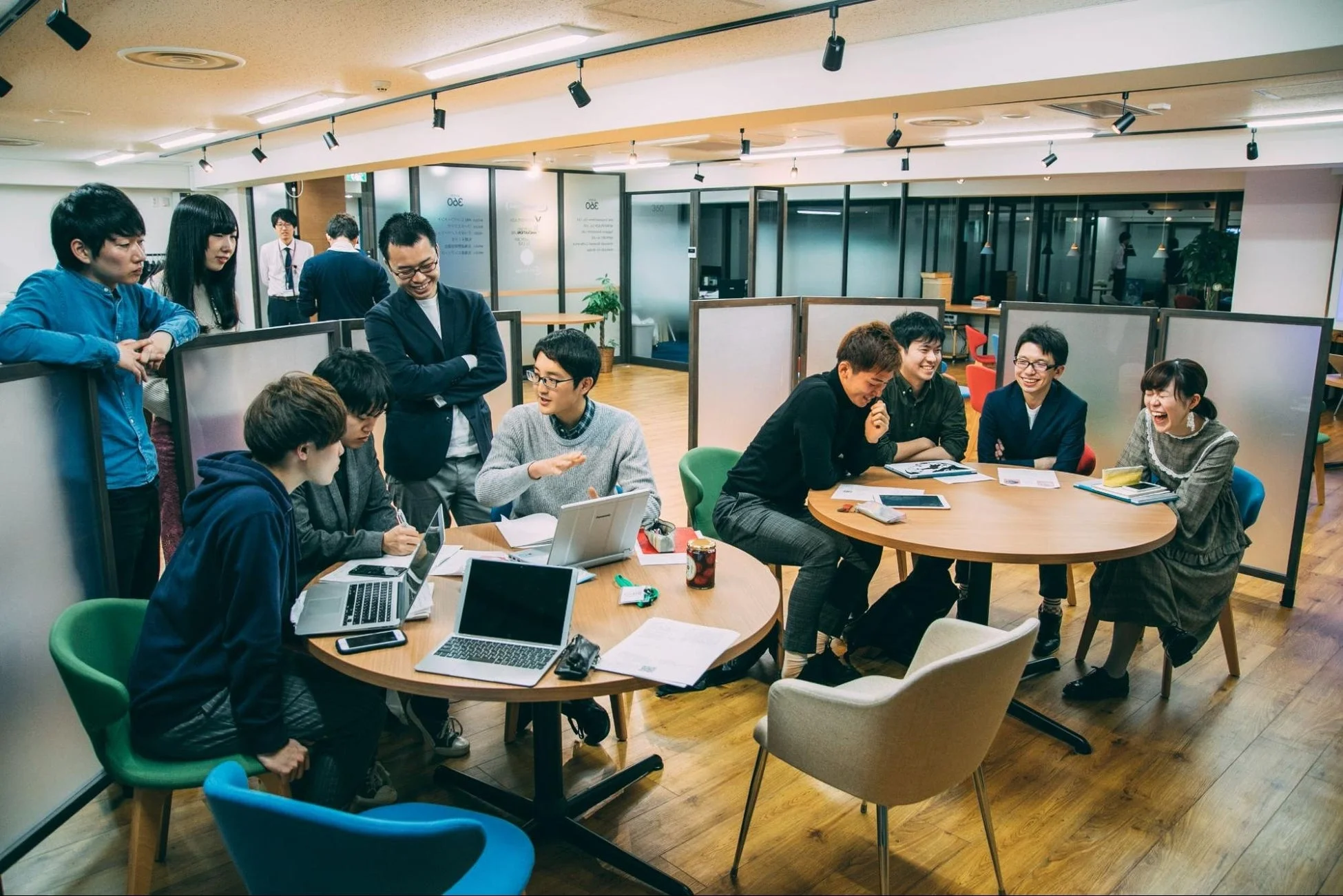 オフィスで複数の若者たちが集まり、笑顔で活発に議論や共同作業をしている様子。