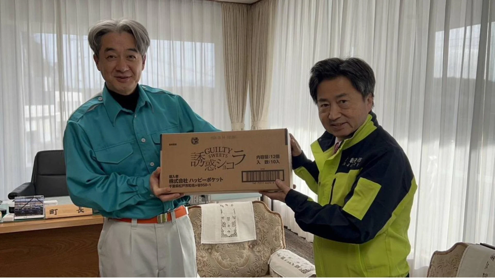 執務室で、町長服を着た男性と人吉市のジャケットを着た男性が、笑顔で「誘惑ショコラ」の箱を受け渡しています。
