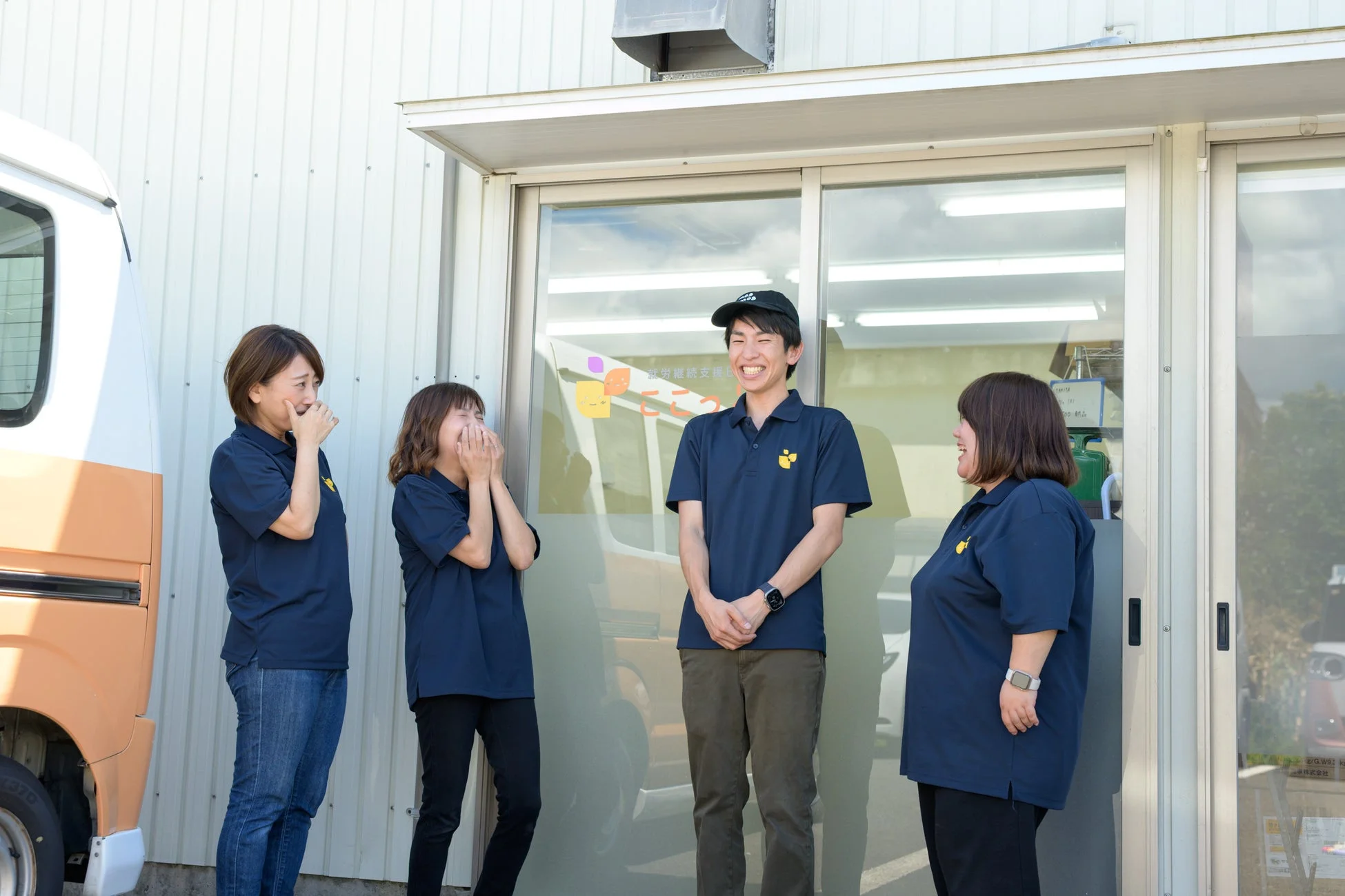 青いポロシャツを着た4人の人々が建物の前で笑顔で談笑している様子。就労継続支援事業所のスタッフの日常風景と見られる。