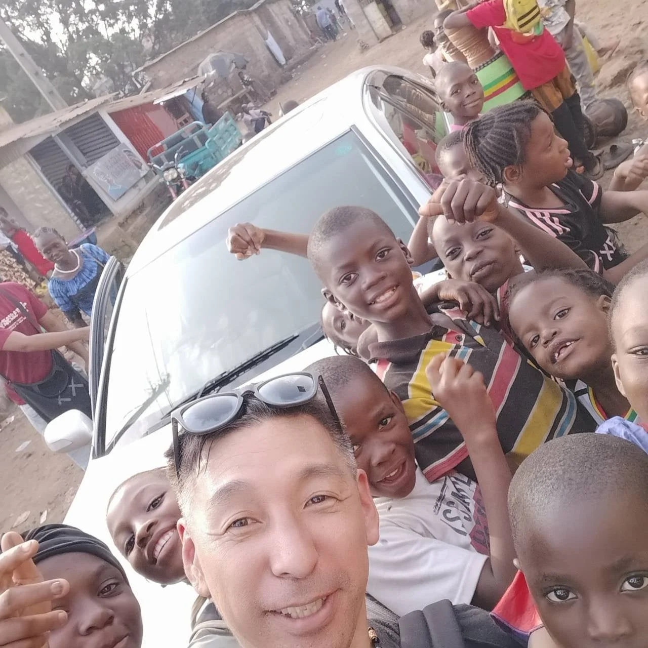 一人の男性が、白い車の周りに集まった多くのアフリカの子供たちと笑顔でセルフィーを撮っています。