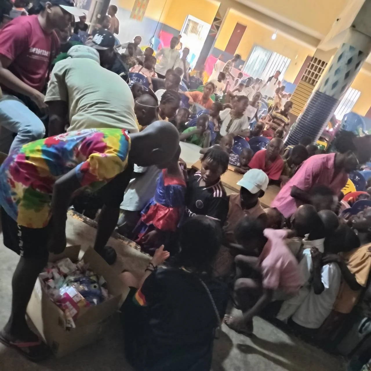 大勢の子供と大人が室内に集まり、大人たちが段ボール箱から物資を配布している様子。