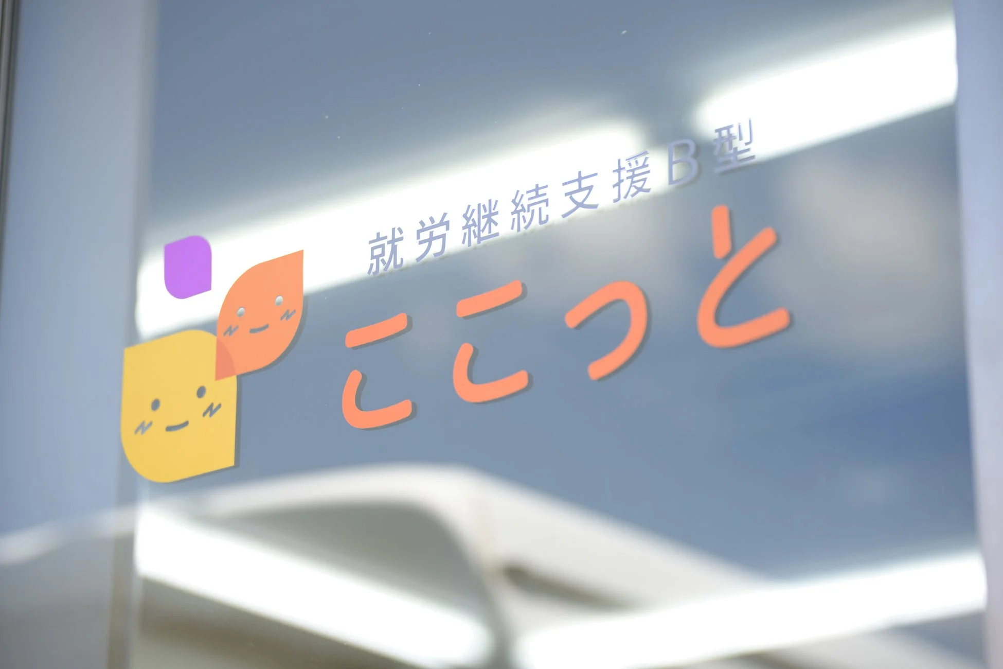 ガラスに「就労継続支援B型 ここっと」という文字と、可愛らしいキャラクターのロゴが貼られています。就労支援施設の入口のサインボードと推測されます。