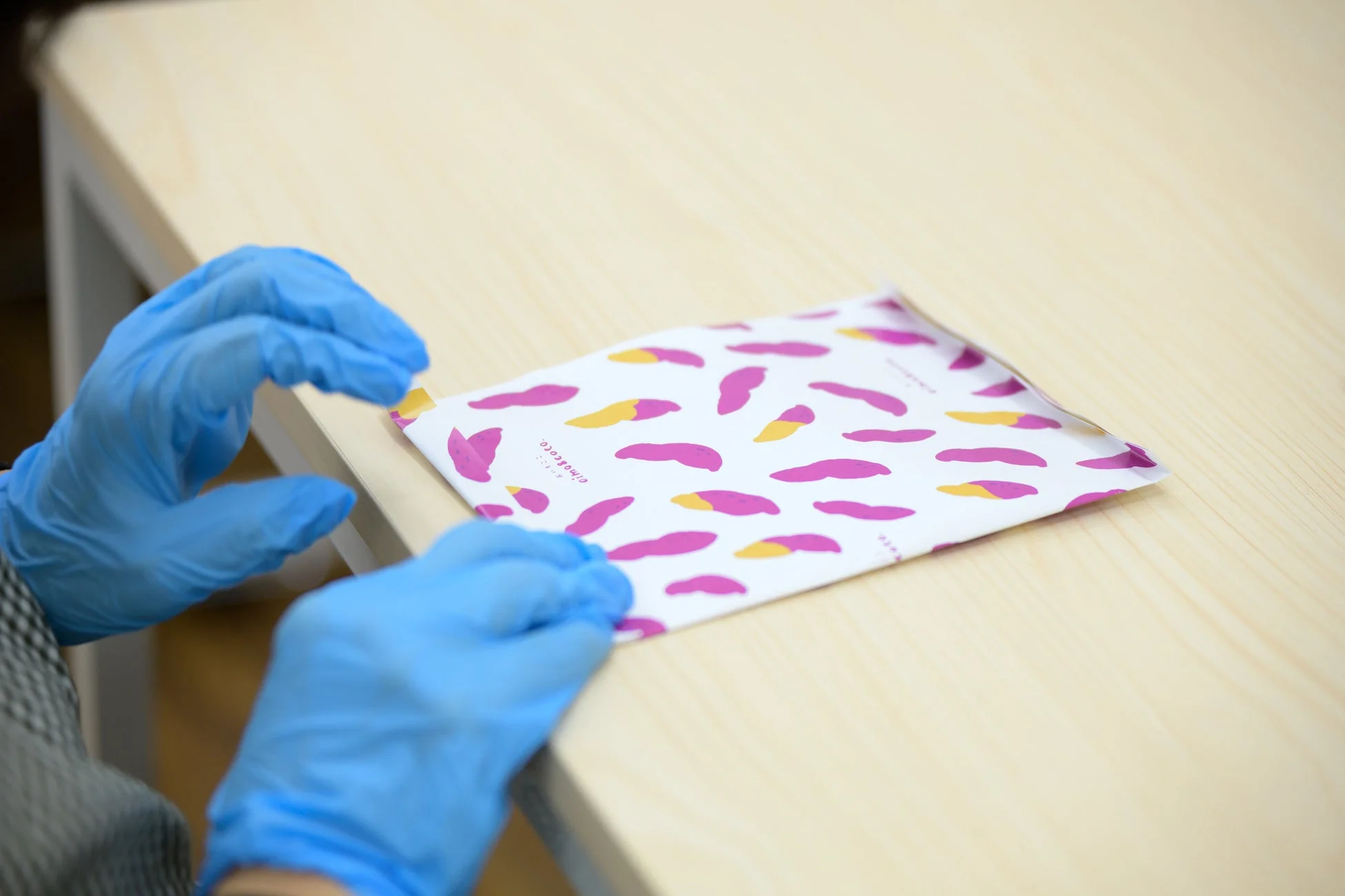 青い手袋をはめた手が、サツマイモのような模様が描かれた白いパッケージを扱っています。食品の準備か梱包作業を行っている様子です。