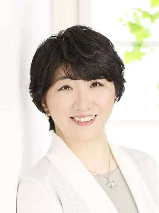 笑顔でカメラを見つめるショートヘアの日本人女性のポートレート。白いカーディガンを羽織り、シンプルなネックレスを身につけており、親しみやすい印象を与えます。