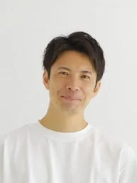 白い背景の前で、白いTシャツを着たアジア系の男性がカメラに向かって優しく微笑んでいます。短く黒い髪で、清潔感のある印象です。