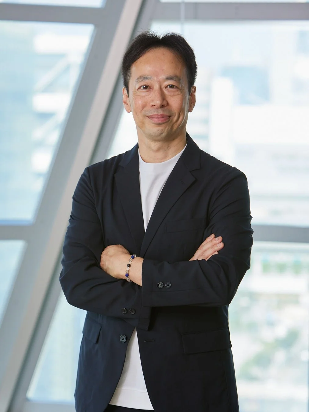 現代的なオフィスを背景に、ダークスーツと白いTシャツを着用した中高年の日本人男性が、腕を組んで穏やかに微笑んでいます。ビジネスシーンや企業の代表者を示すポートレートです。