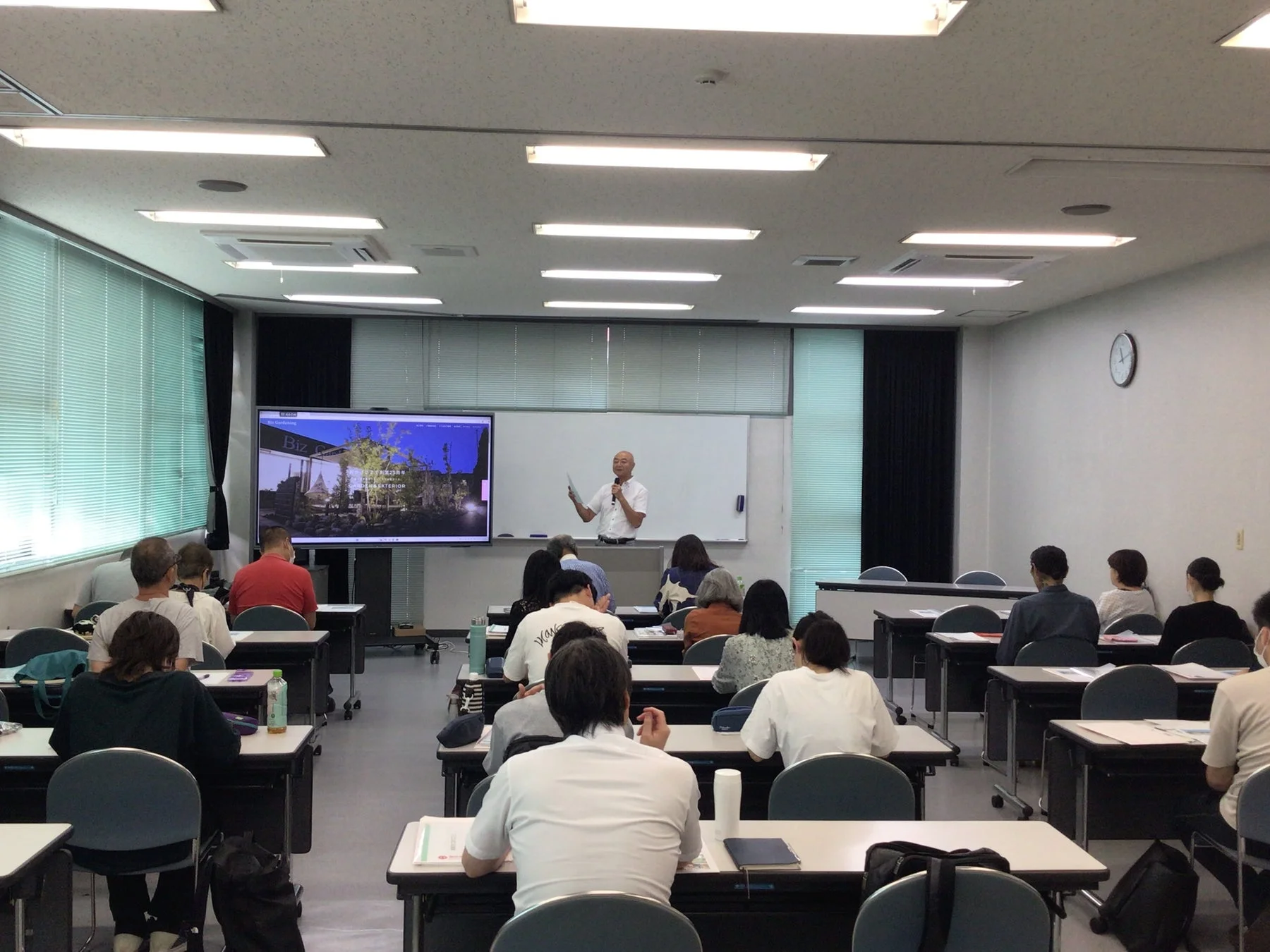 教室で講師がマイクを持ち、プロジェクターに映し出された画面を背景に、多数の受講者に向けて話しているセミナー風景です。