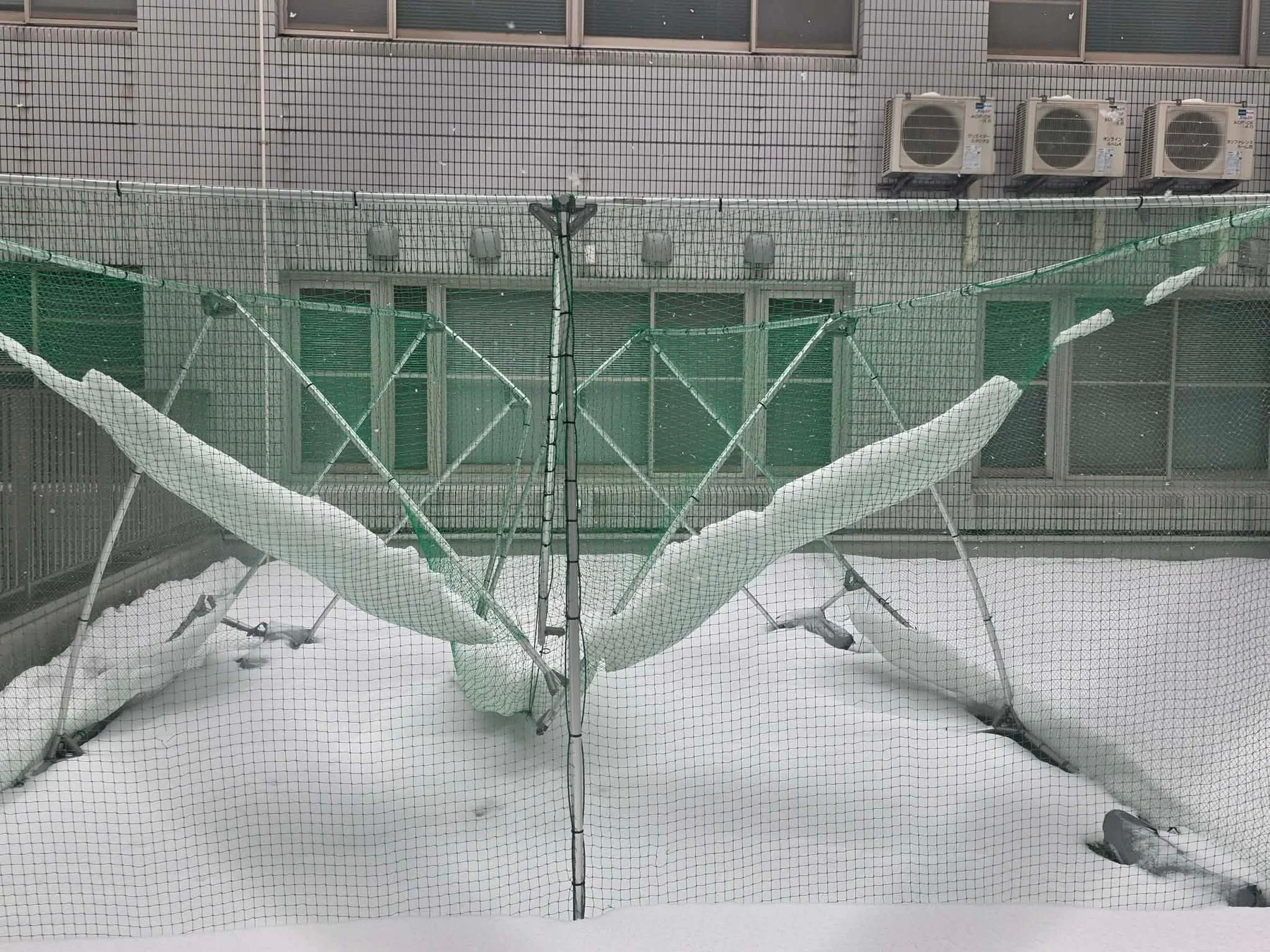 雪が降り積もり、緑色の防球ネットが白い雪で覆われている冬の風景です。ネットの下の地面も一面の雪で、背景には室外機が設置された建物が見えます。
