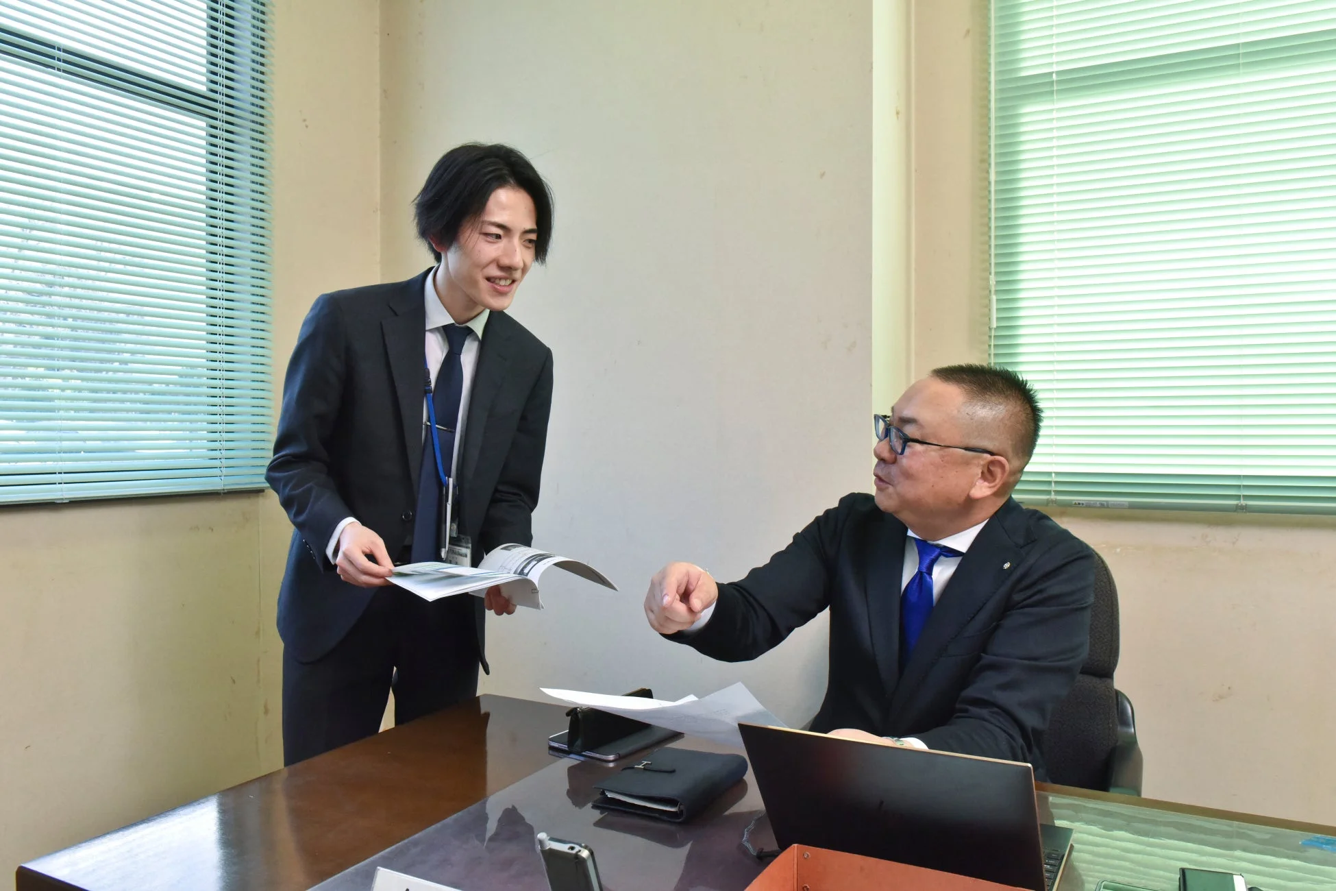 オフィスで二人のビジネスマンが書類を囲んで打ち合わせをしている様子。