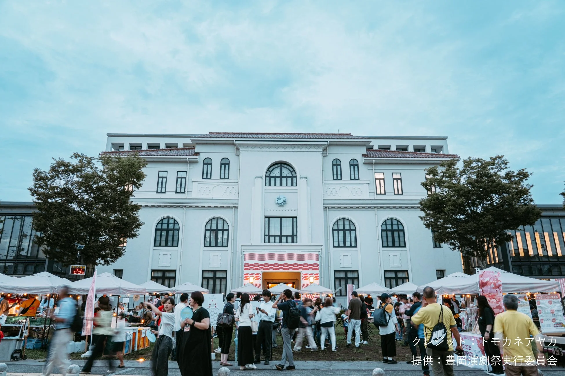 歴史ある白い建物の前で、多くの人々が賑わう屋外イベントの様子。屋台やテントが立ち並び、夕暮れ時の活気ある雰囲気が伝わる。豊岡演劇祭に関連する催しと思われる。