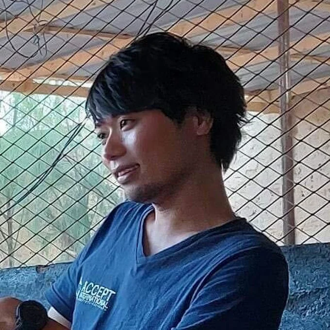 黒い短髪の若いアジア人男性が青いTシャツを着て、金網の前で横を向いているポートレート