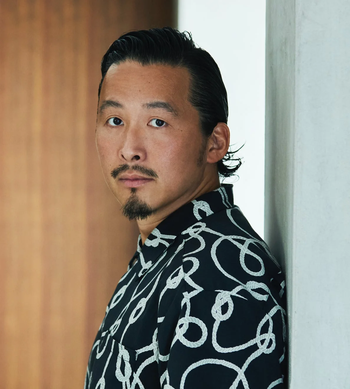 黒いロープ柄のシャツを着た男性のポートレート。真剣な表情でカメラを見つめており、背景には木製の壁と白い壁が見える。髭とオールバックの髪型が特徴的。