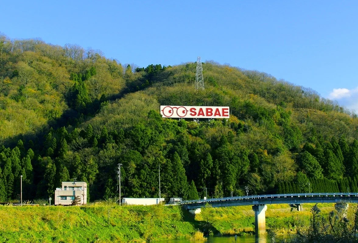 福井県鯖江市のランドマークである「SABAE」の巨大な看板が緑豊かな山腹に設置されている風景。青空の下、手前には川と橋が架かり、のどかな日本の景色が広がっている。