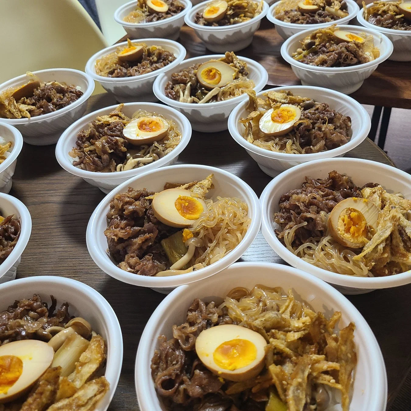 テーブルに、牛肉、卵、しらたき、きのこなどが盛り付けられた丼物が多数の白い容器に入って並べられています。