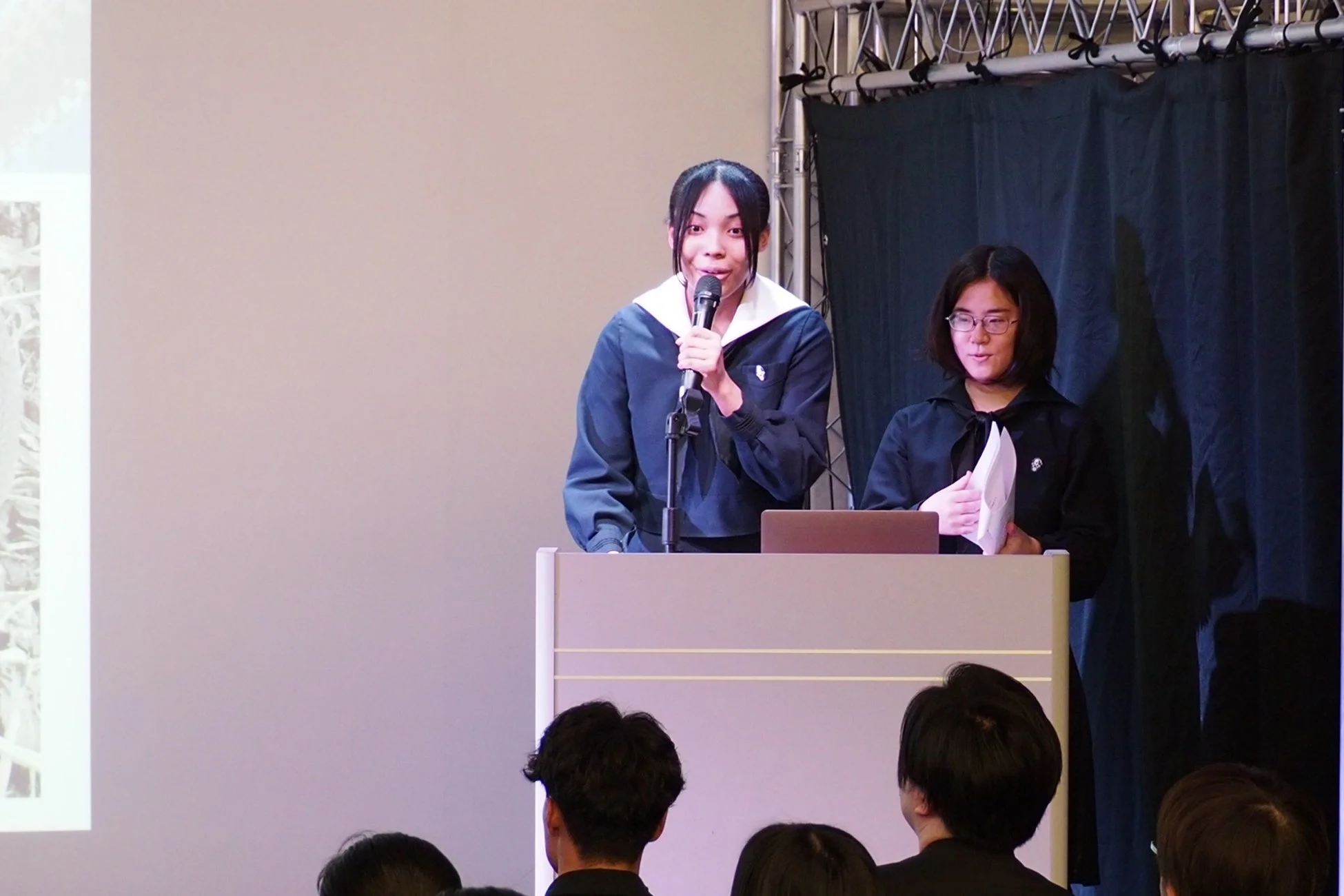 制服を着た2人の若い女性が壇上に立ち、一人がマイクを持って話しています