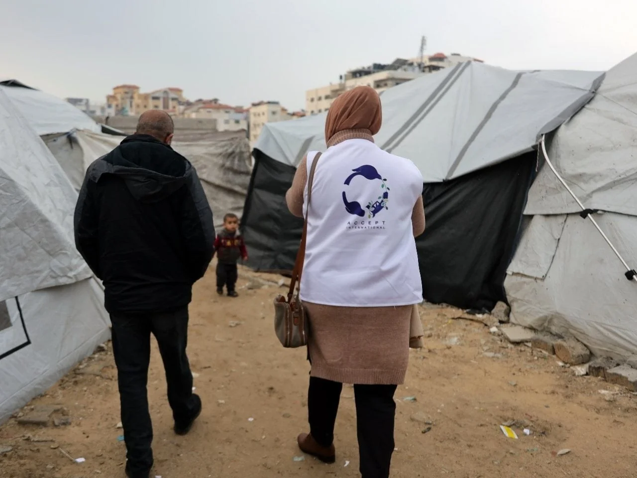 難民キャンプを歩く人々。一人の大人は「ACCEPT INTERNATIONAL」と書かれたベストを着用