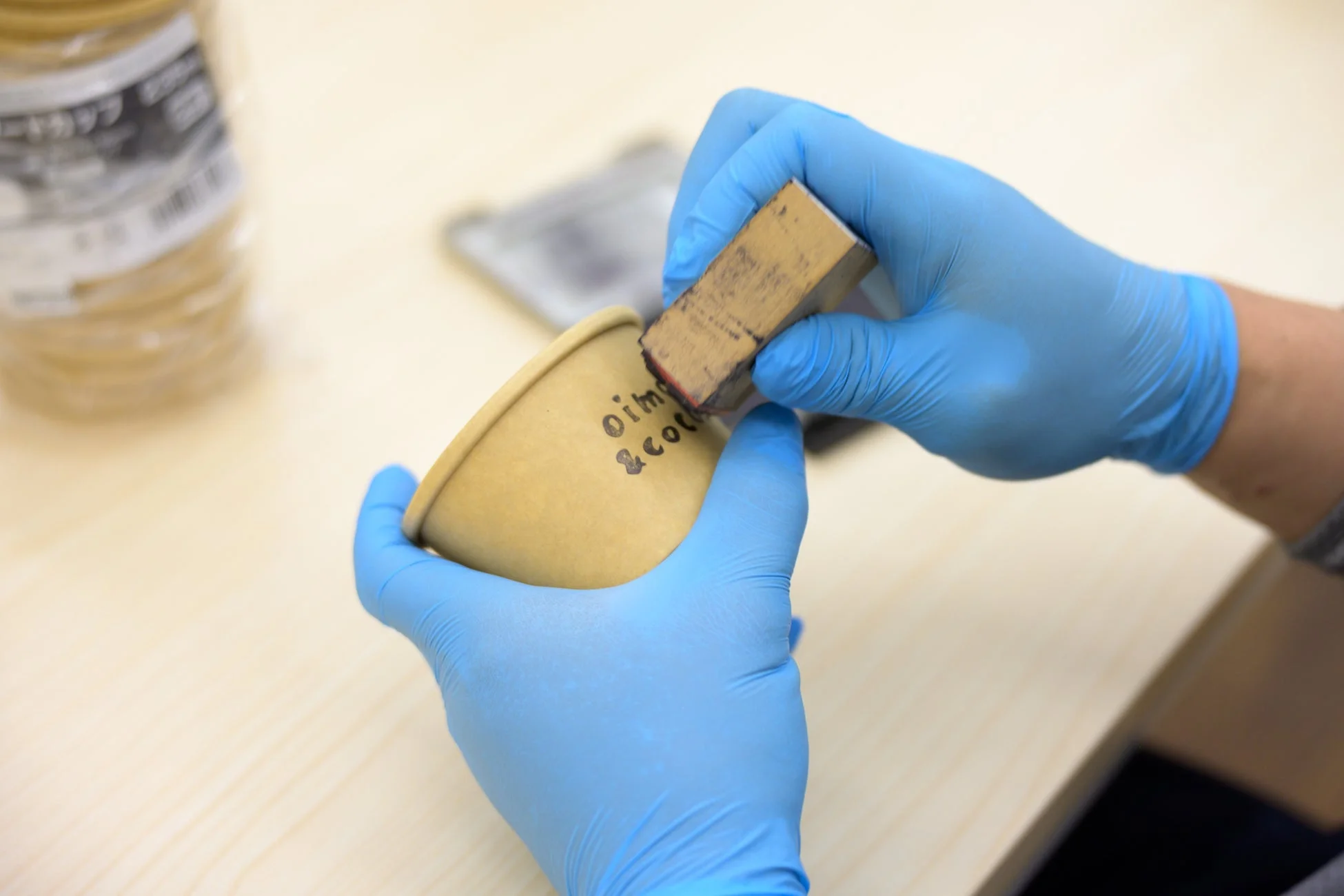 青い手袋をはめた手が、紙コップに「oim & cotf」という文字のロゴをスタンプで押している様子。オリジナルカップを製作する手作業の工程を捉えた画像です。