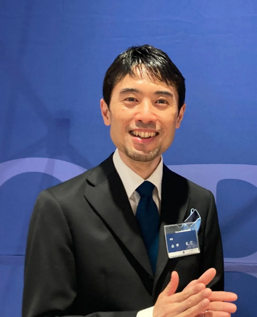 青い背景の前で笑顔を見せるビジネスマンの男性。黒いスーツに青いネクタイを着用し、胸元には「山中祐二」と書かれた名札をつけています。会議やイベントの一場面のようです。