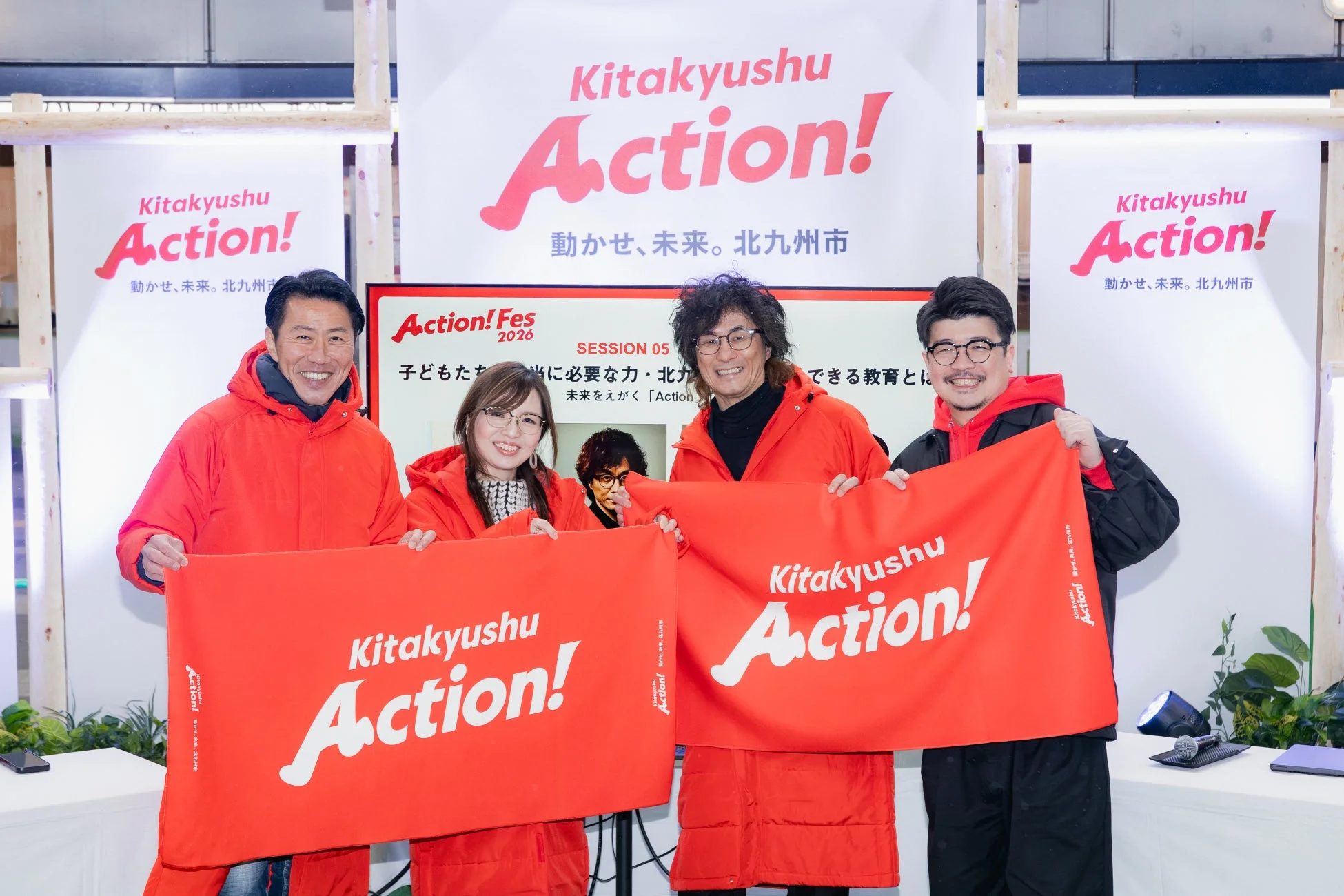 「Kitakyushu Action! Fes 2026」イベントで、赤い服を着た4人が「Kitakyushu Action!」バナーを掲げ笑顔で集合写真