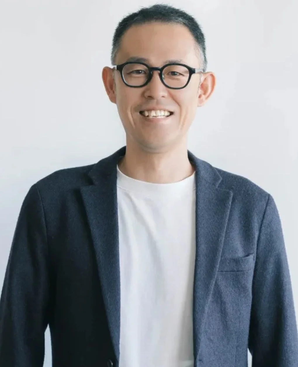 白い背景の前で、メガネをかけ紺色のブレザーと白いTシャツを着た男性が、穏やかな笑顔を見せている上半身のポートレート写真です。