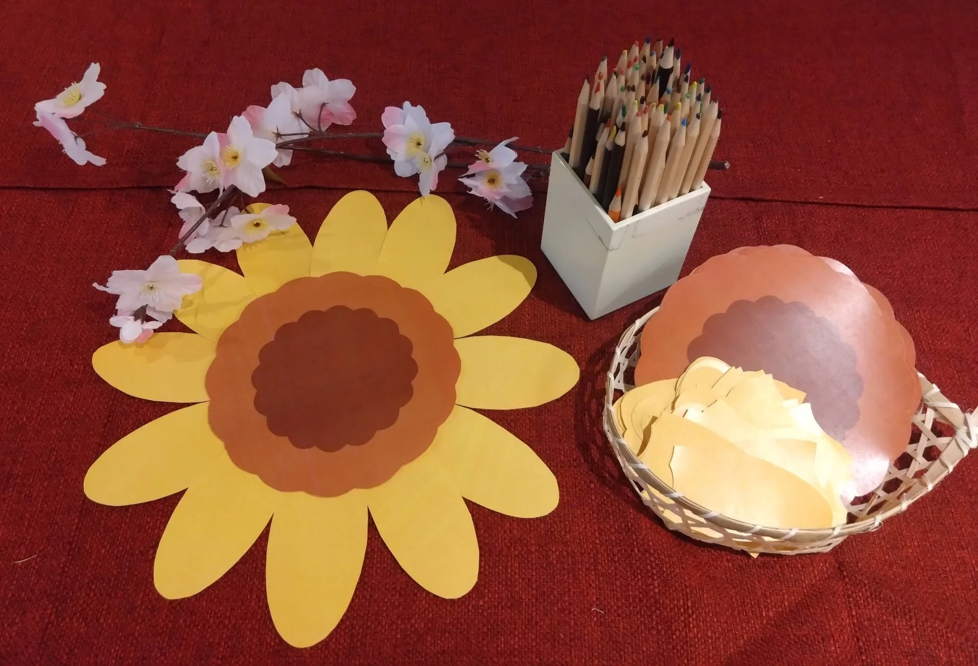 赤い布の上に、紙製のひまわり、桜の造花、色鉛筆、そしてバスケットに入った紙の材料が並べられています