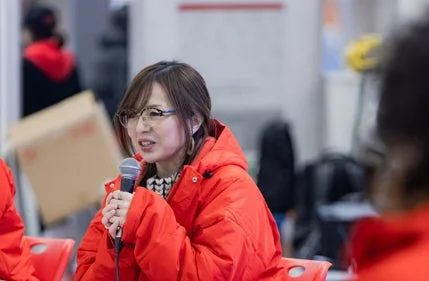 赤いジャケットを着た女性がマイクを手に持ち、真剣な表情で話している様子