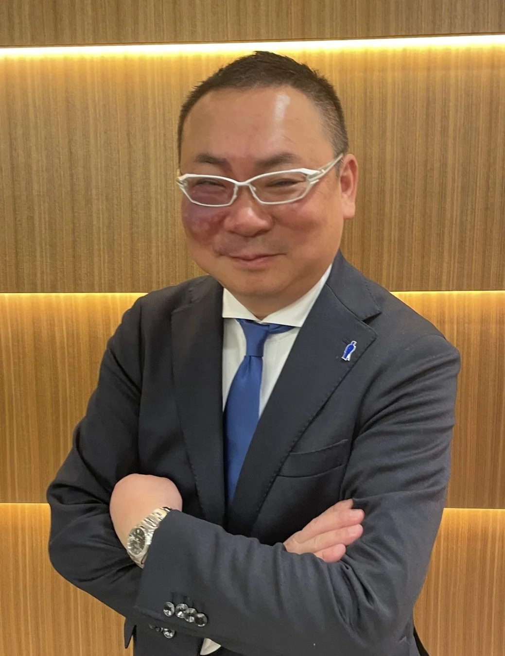 スーツを着て眼鏡をかけた男性が、木目調の壁を背景に腕を組み、穏やかに微笑んでいるポートレート写真です。