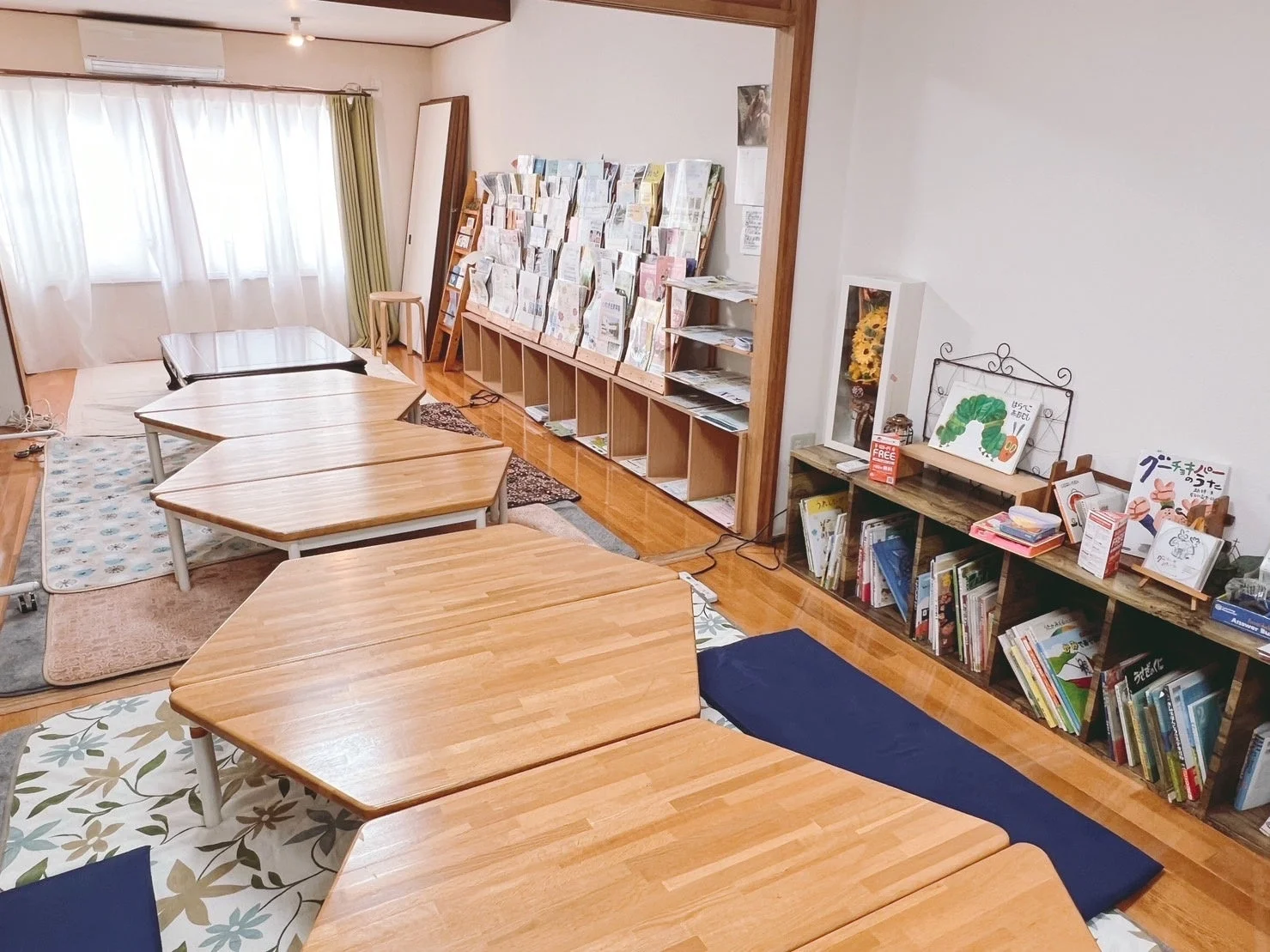 明るく居心地の良い部屋で、複数の低い木製テーブルが並べられています。壁際には多くの本、特に絵本が並べられた本棚があり、子供向けの読書や学習スペースとして使われているようです。