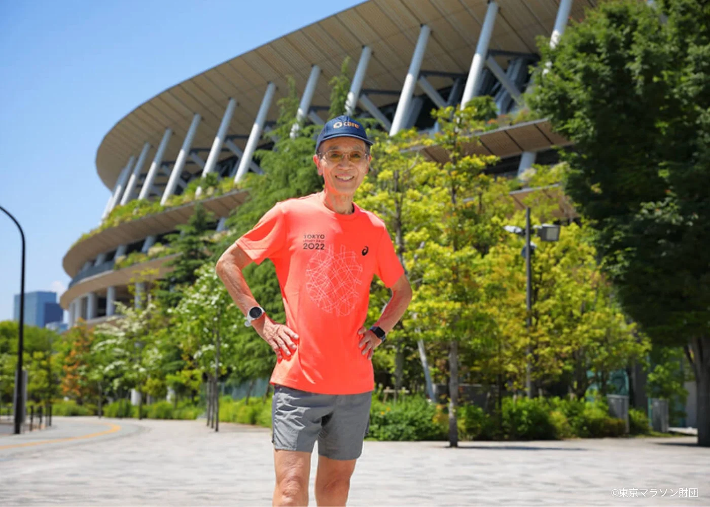晴れた青空の下、ランニングウェアを着用した男性が国立競技場を背景に笑顔で立っている。オレンジ色のTシャツには「TOKYO 2022」と書かれており、東京でのスポーツイベント、特にマラソンに関連する様子が伺える。健康的なライフスタイルと活動的な雰囲気が伝わる一枚。