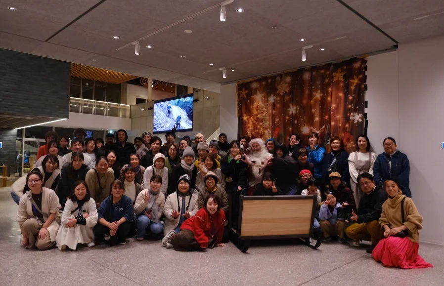 サンタクロースを囲んで大勢の人々が笑顔で集合写真を撮っている屋内イベントの様子