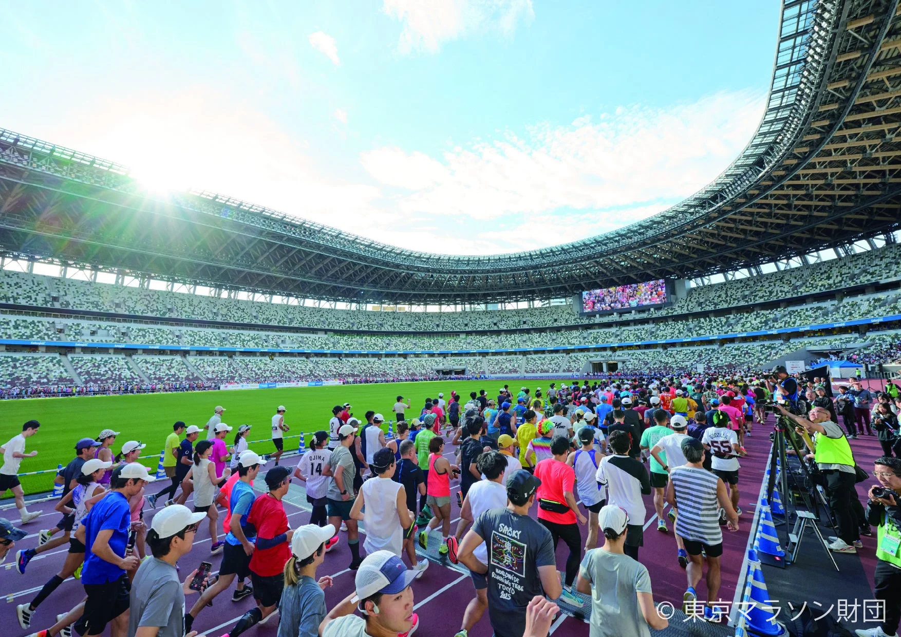 晴れた青空の下、多くの人々が広大な競技場のトラックを走る大規模なランニングイベントの様子。参加者たちが活気ある光景を繰り広げている。