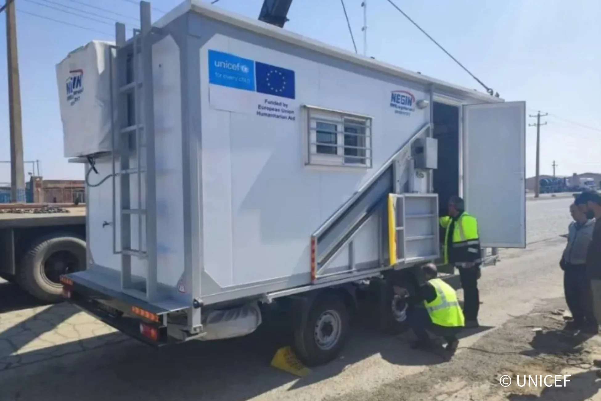 UNICEFと欧州連合による人道支援を受けた移動式ユニットがトラックに積載され、作業員がその設置や点検を行っている様子。青空の下、屋外での支援活動の一場面を捉えています。
