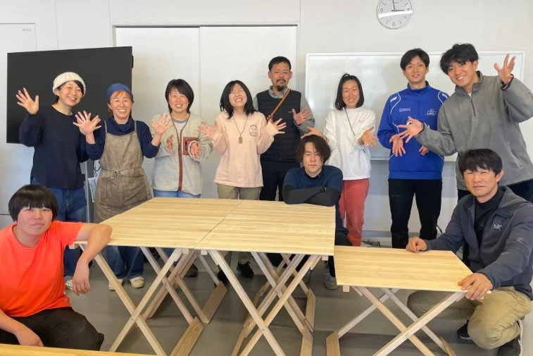 明るい室内で、笑顔の大人たちが木製の折りたたみテーブルを囲んで集合写真を撮っています