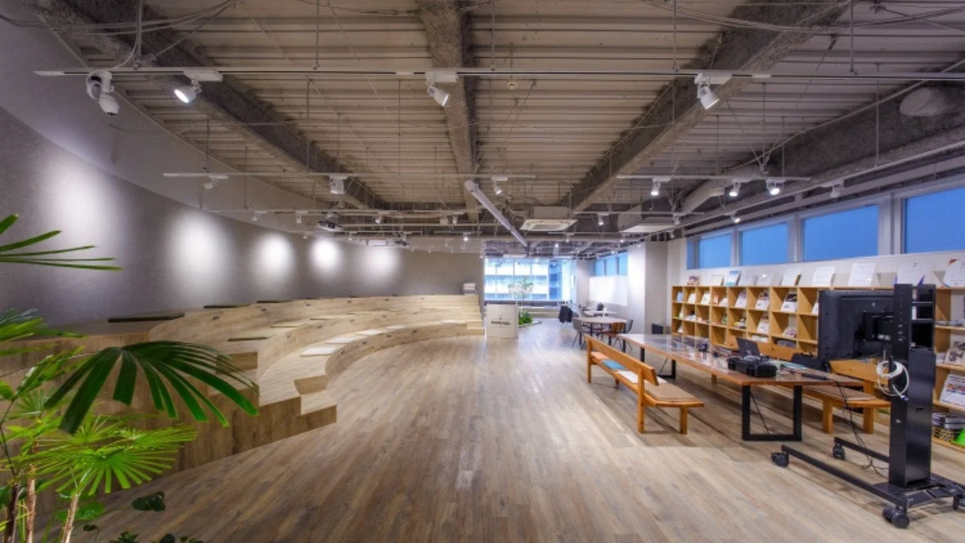 モダンで開放的なオフィスまたはコワーキングスペース。木製の階段状の座席エリア、長い作業テーブル、本棚があり、多様な働き方に対応した空間です。