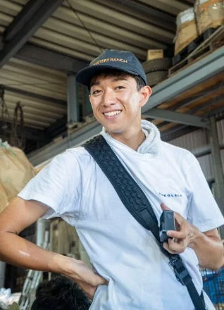 笑顔の若い男性が白いTシャツとキャップを着用し、肩にカメラを提げています