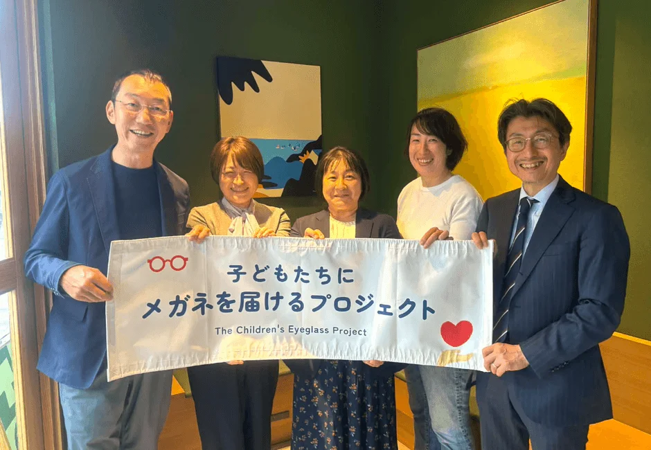 子どもたちにメガネを届けるプロジェクトのメンバー5人が、笑顔でバナーを掲げている集合写真です。