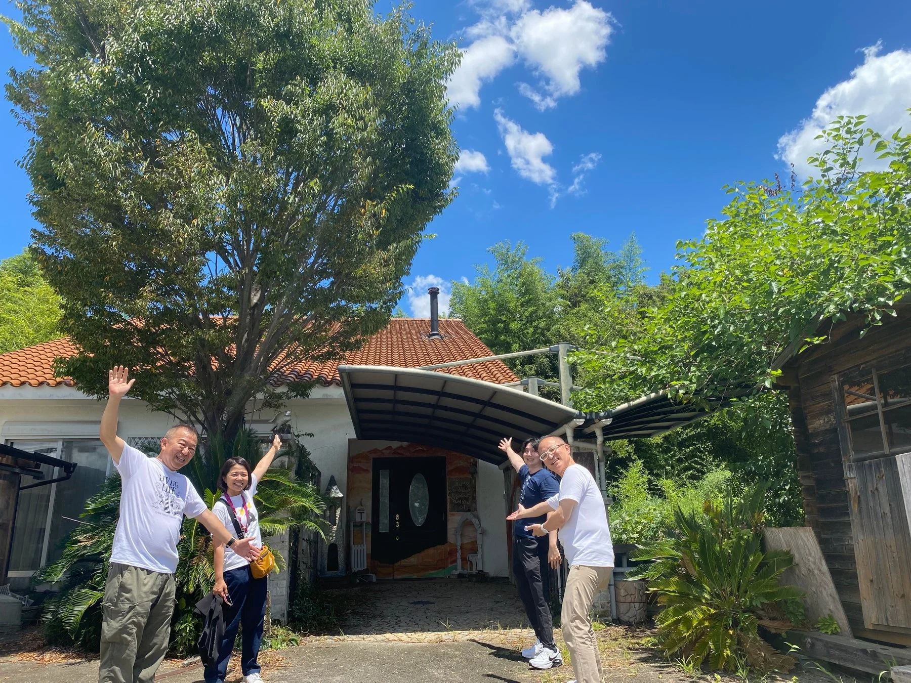 晴れた日に、タイル屋根の家と豊かな緑を背景に、4人の男女が笑顔で手を振っている様子。